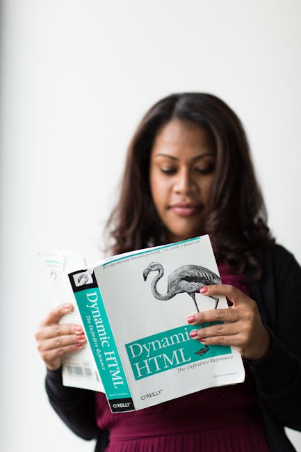 A woman with dark brown hair, dressed in a black blazer over a burgundy top, is reading a book titled 'Dynamic HTML' with a bird illustration on the cover. She is standing against a plain white backgr